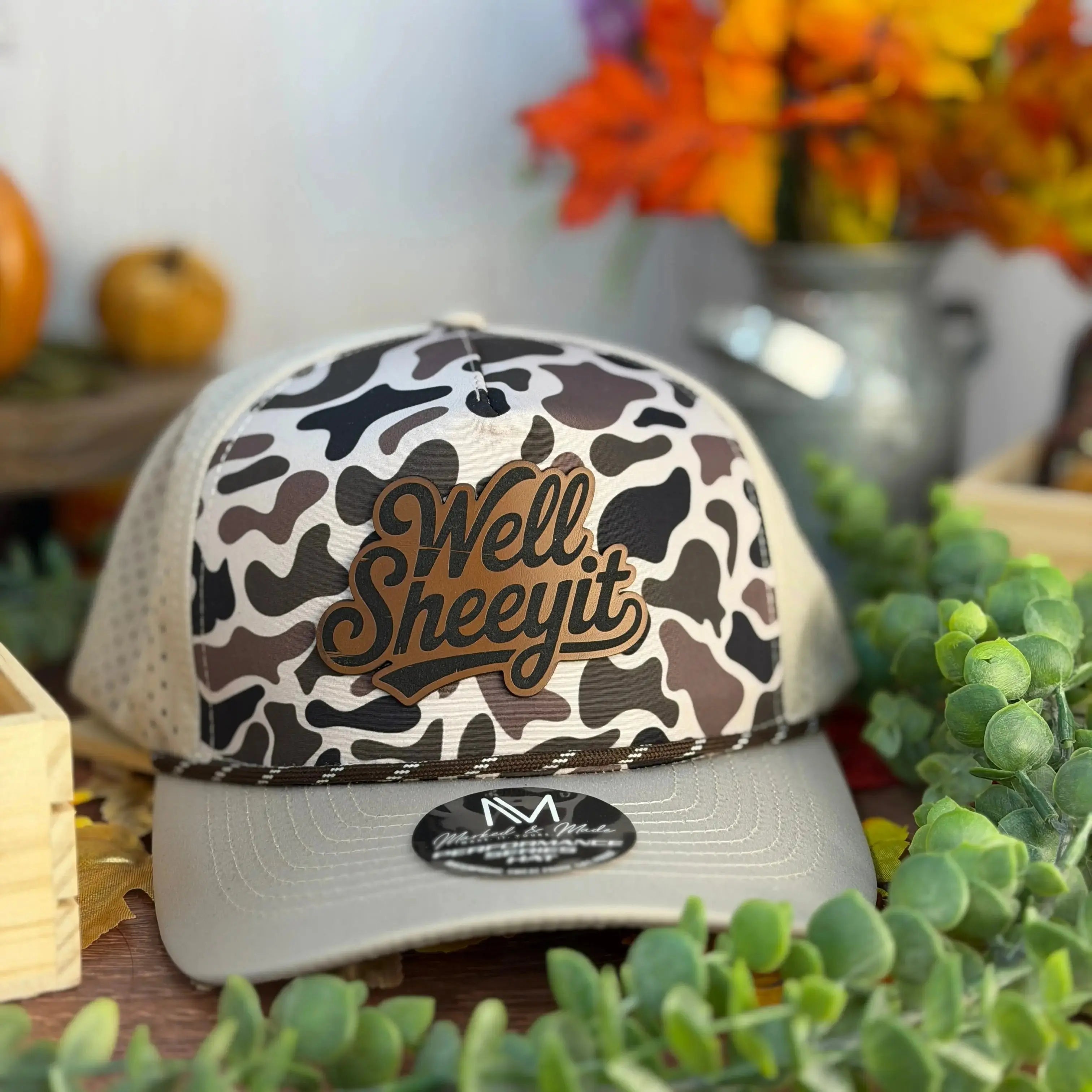 A camouflage-patterned baseball cap with the text "Well Sheety It" printed on it, placed on a table surrounded by various objects such as a vase of flowers, a wooden crate, and a potted plant.