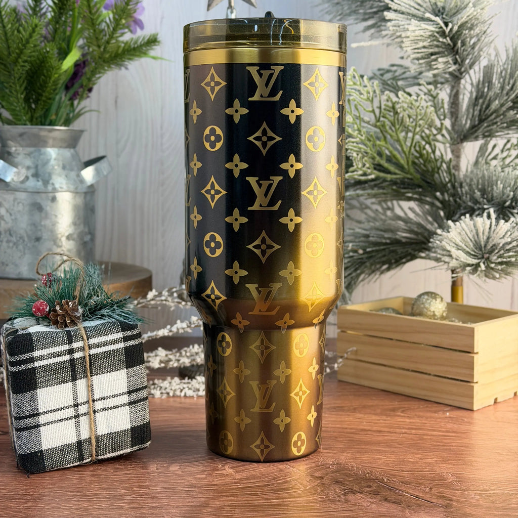 a large, gold-colored tumbler with a black and gold pattern, placed on a wooden surface. In the background, there are some decorative elements such as a small Christmas tree and a gift box.