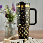 A black and gold patterned thermos mug with a handle, placed on a wooden surface next to a vase of purple flowers.