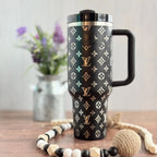 A black and white patterned thermos mug with a handle, placed on a wooden surface alongside a beaded necklace and a vase of purple flowers.