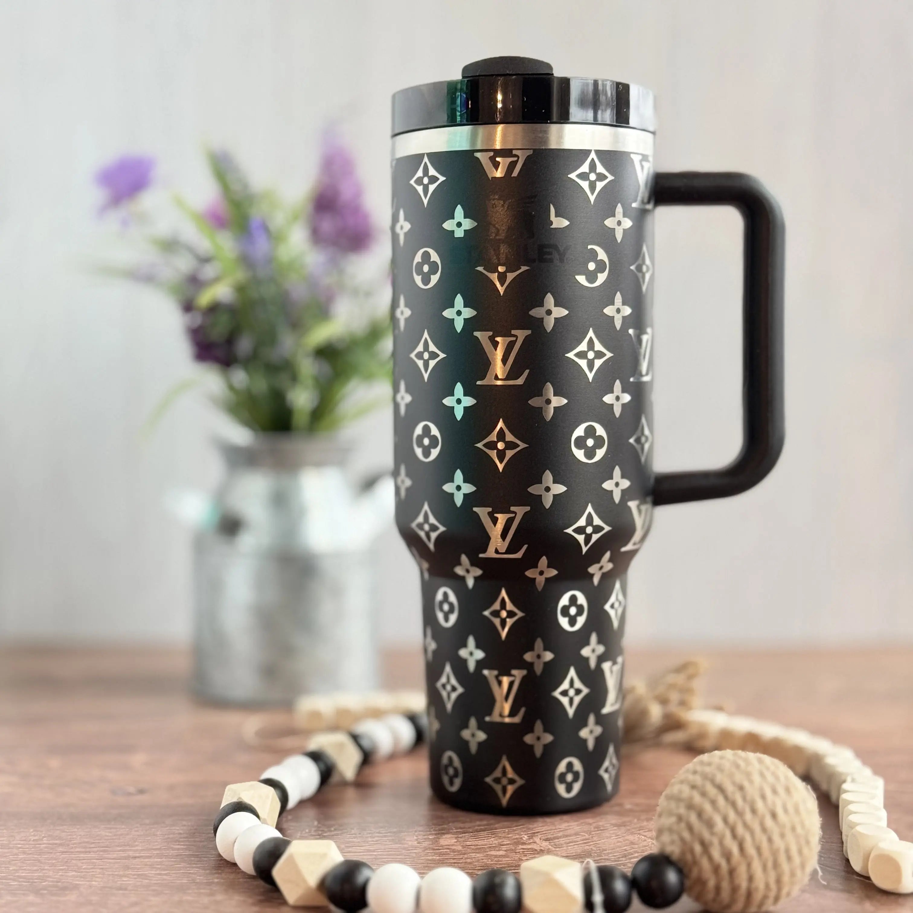A black and white patterned thermos mug with a handle, placed on a wooden surface alongside a beaded necklace and a vase of purple flowers.