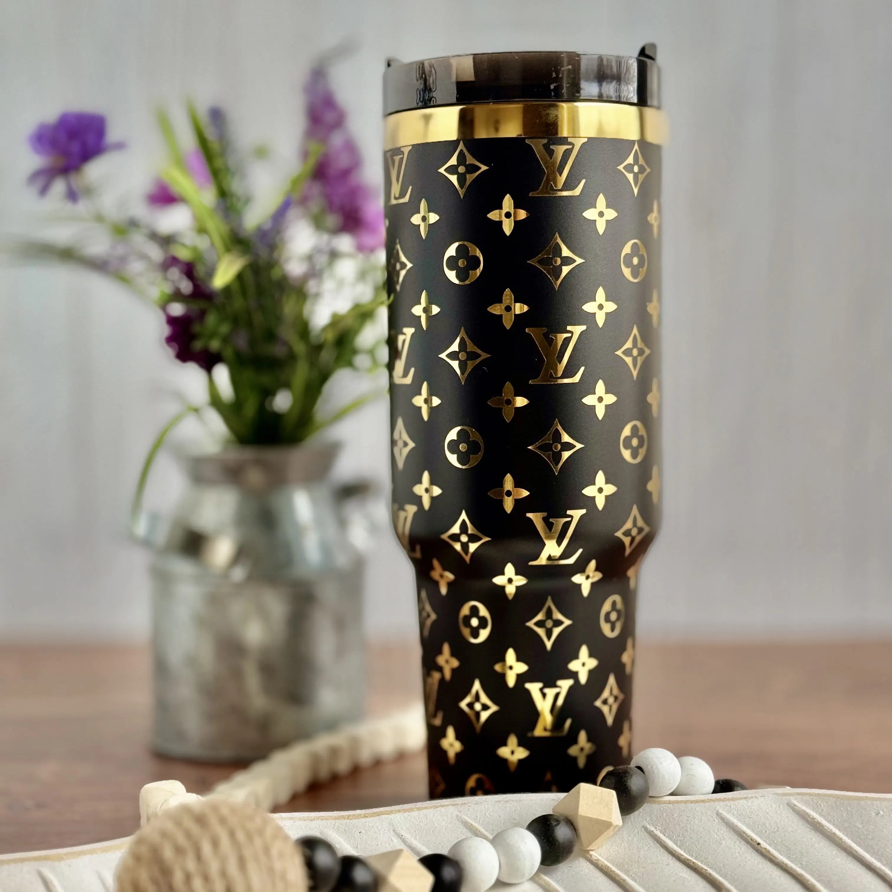 a black and gold Louis Vuitton-patterned tumbler mug placed on a wooden surface, with a vase of purple flowers in the background.