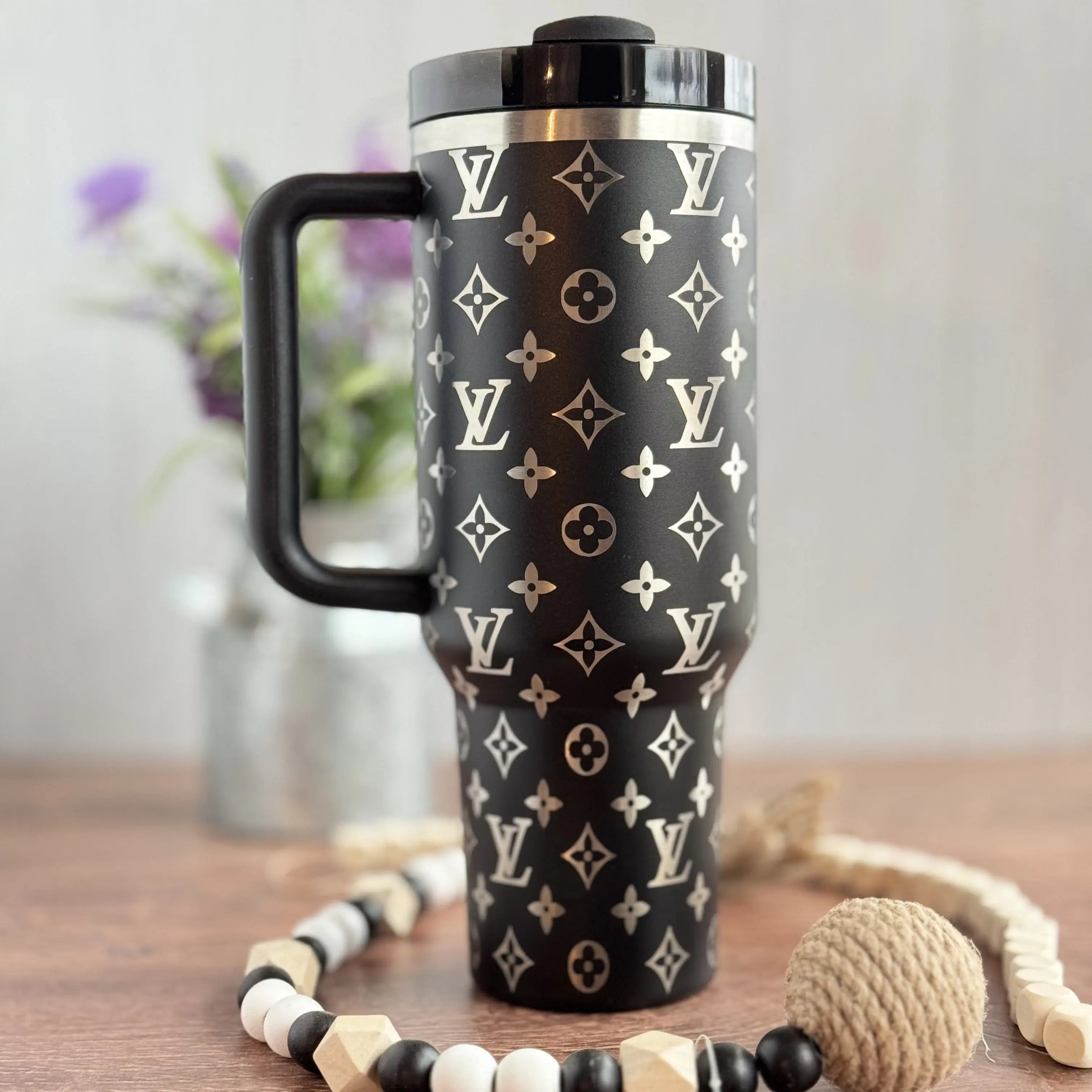 A black and white patterned thermos mug with a handle, placed on a wooden surface alongside a beaded necklace and a small ball.