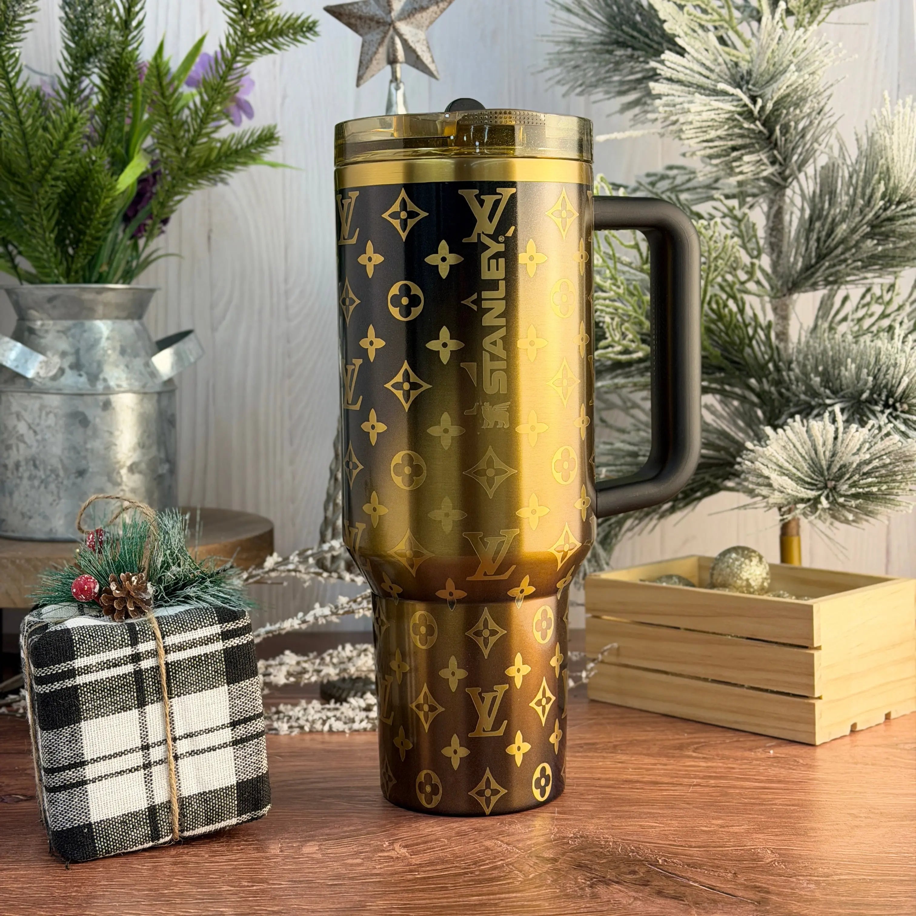 a large, gold-colored thermos mug with a black handle, placed on a wooden surface. In the background, there is a Christmas tree and some decorative items, creating a festive atmosphere.