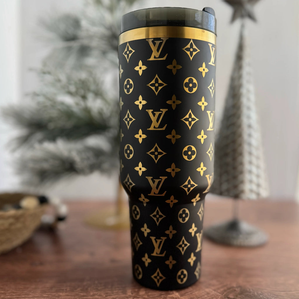 A black and gold patterned thermos with a gold-colored lid, featuring the Louis Vuitton monogram logo, is displayed on a wooden surface.